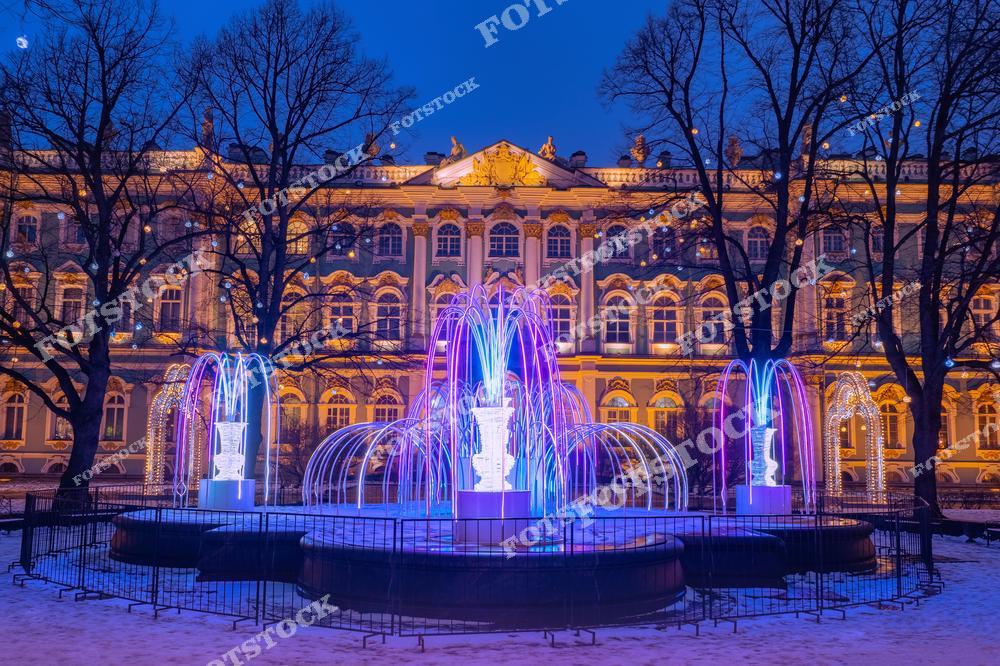 На фотографии изображены фонтаны, украшенные подсветкой в вечернее время. За ними видно здание Эрмитажа (Зимнего дворца) в Санкт-Петербурге. Эти декорации создают праздничное настроение, они украшают город к Новому году или Рождеству. Такое фото могут использовать для туристических проспектов, в качестве рекламы городских зимних развлечений или для иллюстрации статей о зимних праздниках в России.